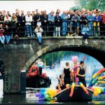 Boat passing under a bridge while people watch from above.